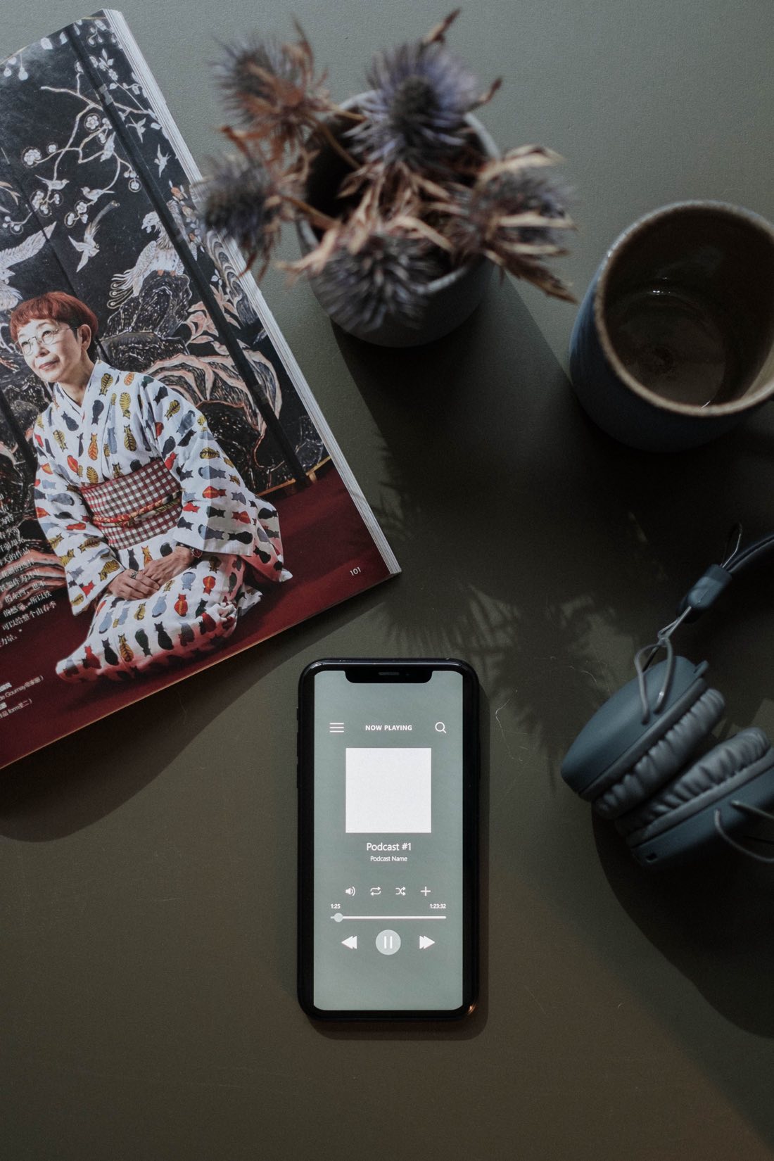 A phone playing a podcast on a desk, beside headphones, a coffee cup and an open magazine.